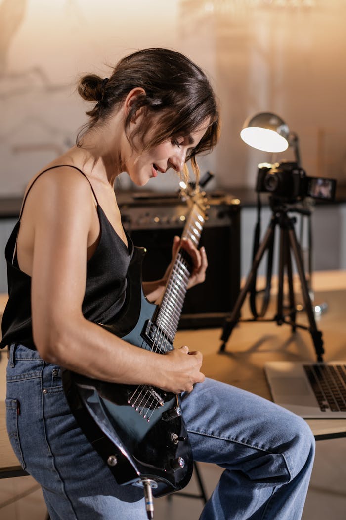 A woman playing an electric guitar indoors with recording equipment set up nearby.